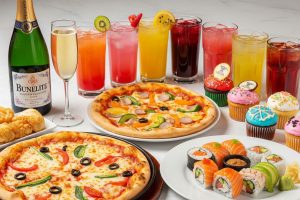 A vibrant spread of diverse food and drinks, including two pizzas, sushi rolls, cupcakes, and various colorful beverages in glasses, with a bottle of sparkling wine.